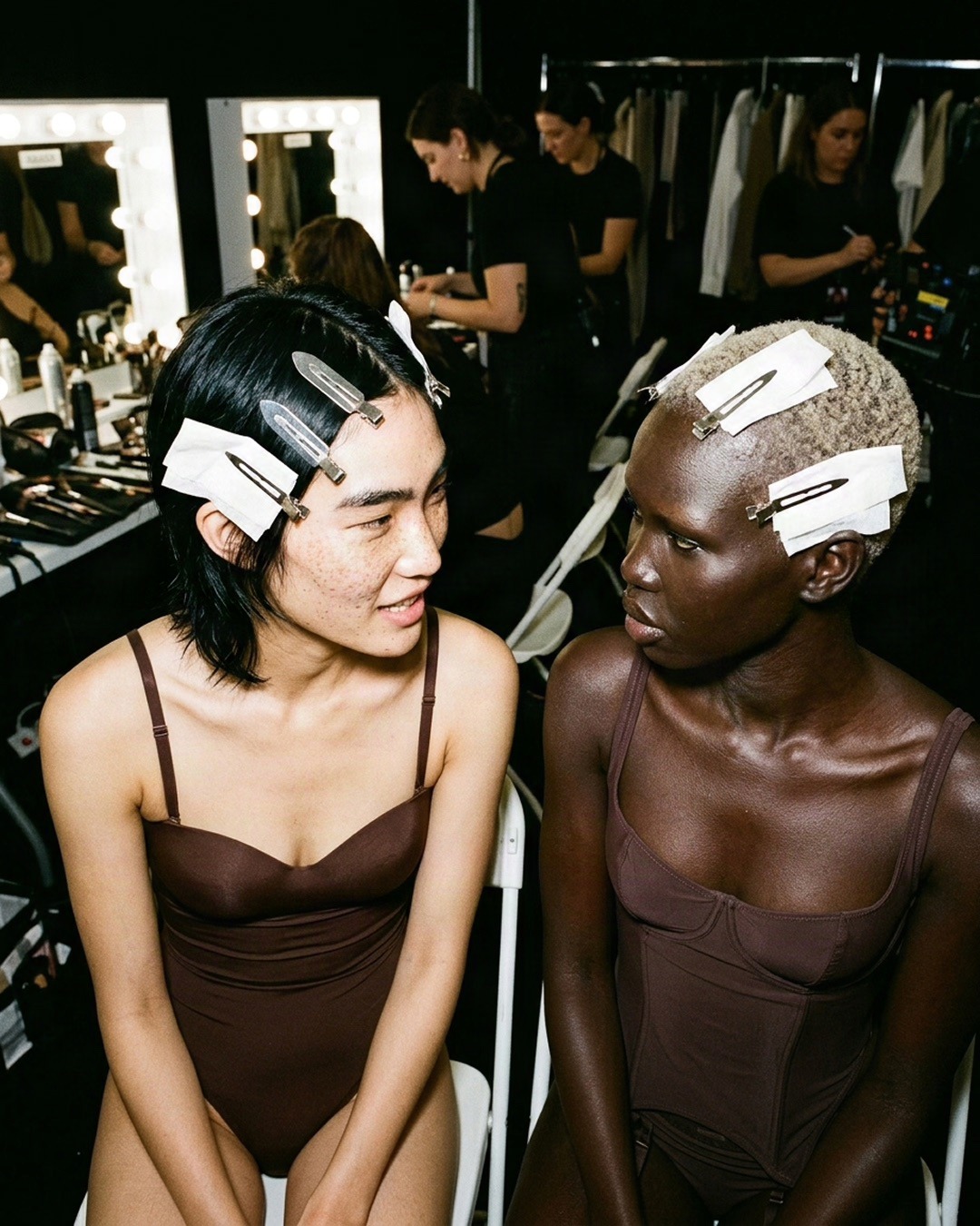 SKIMS spec editorial portrait of two models in brown bodywear facing each other backstage with clipped hair