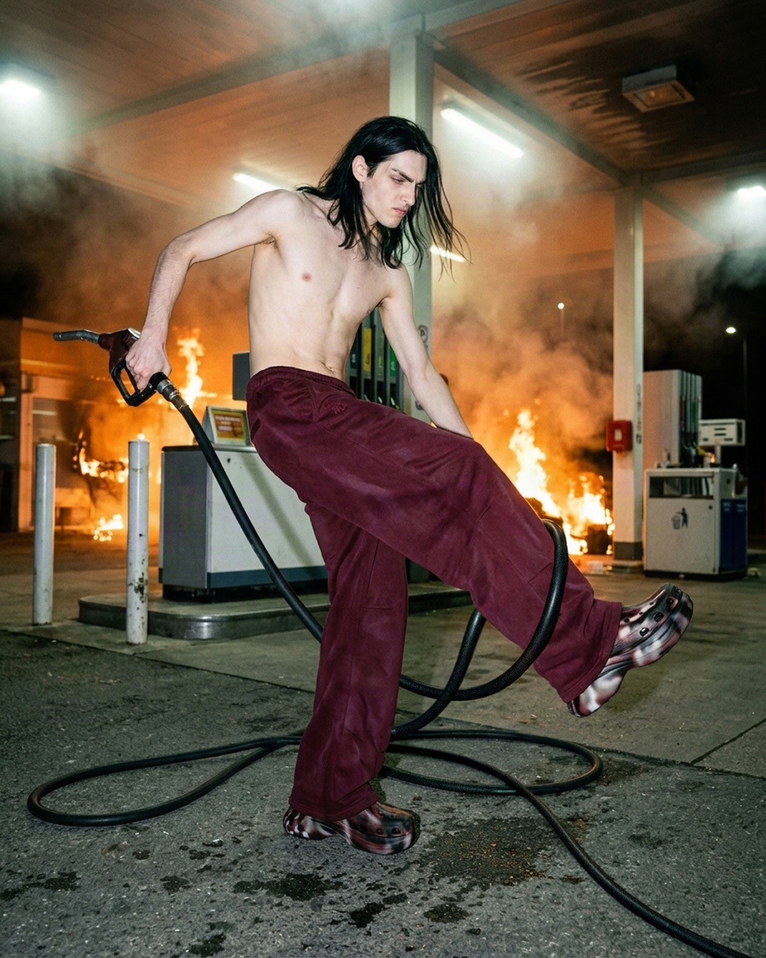 Mowalola spec editorial model posing with a fuel nozzle and burgundy trousers against orange flames